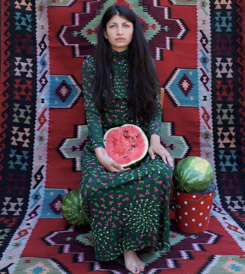slika sa izložbe na kojoj sedi žena crne duge kose na tapaciranoj šarenoj pozadini i u krilu drži polovinu lubenice