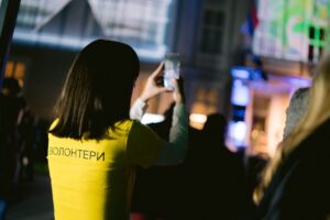 volonteri na događaju u gradu, devojka u žutoj majici okrenuta leđima fotografiše u gradu putem mobilnog